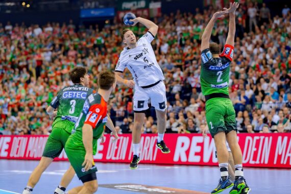 Ein Handballspieler in einem weißen Trikot springt, um den Ball in Richtung Tor zu schießen, während drei Gegner in grünen Trikots versuchen, ihn während eines Hallenspiels vor einer großen Zuschauermenge zu blockieren.