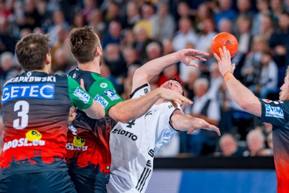 Drei männliche Handballspieler in roten, schwarzen und weißen Trikots kämpfen um einen orangefarbenen Ball. Der Spieler in Weiß greift nach hinten, während zwei Spieler in Rot und Schwarz ihn verteidigen. Im Hintergrund ist eine unscharfe Menschenmenge zu sehen.