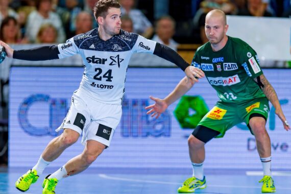 Zwei männliche Handballspieler treten in einer Halle gegeneinander an; der Spieler in Weiß dribbelt den Ball, während der Spieler in Grün den Ball eng verteidigt. Im Hintergrund ist eine unscharfe Menschenmenge zu sehen.