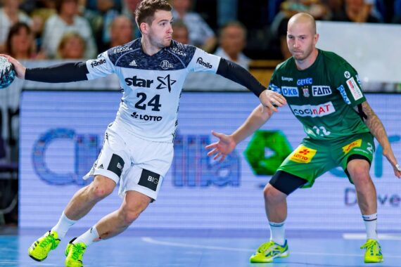 Ein Handballspieler in einem weiß-marinefarbenen Trikot bereitet sich darauf vor, den Ball zu werfen, während er von einem Gegner in einem grünen Trikot während eines Spiels auf einem Hallenplatz eng bewacht wird. Im Hintergrund sind Zuschauer zu sehen.