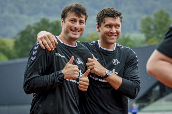 Zwei Männer in passenden schwarzen Sporthemden und Uhren stehen draußen im Regen, lächeln und zeigen die Daumen nach oben. Einer hat den Arm um den anderen gelegt. Grüne Hügel und Bäume sind im Hintergrund verschwommen.