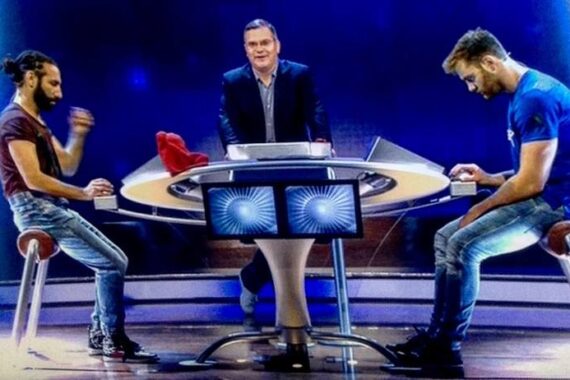 Zwei Männer sitzen sich auf Hockern an einem runden Tisch mit Bildschirmen gegenüber, während ein Moderator hinter dem Tisch steht. Sie befinden sich in einem hell beleuchteten Studio, das an eine Spielshow oder ein Quiz erinnert.