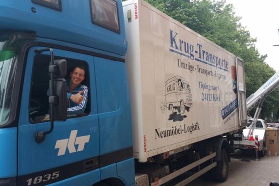 Ein lächelnder Mann sitzt auf dem Fahrersitz eines blauen Umzugswagens von Krug-Transporte und winkt aus dem Fenster. Eine weitere Person steht in der Nähe auf der Straße. Der Lkw ist beschildert und wird in der Nähe von Bäumen und Gebäuden geparkt.