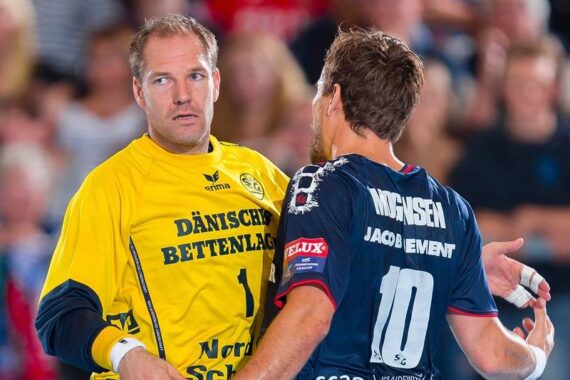 Ein Handballtorwart in einem gelben Trikot steht während eines Spiels einem Spieler in einem dunkelblauen Trikot mit der 10 auf dem Rücken gegenüber, wobei beide auf dem Spielfeld zu kommunizieren scheinen.