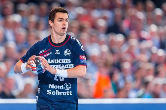 Ein männlicher Handballspieler in einem marineblauen Trikot hält einen Ball während eines Spiels, mit einer unscharfen Menschenmenge im Hintergrund.