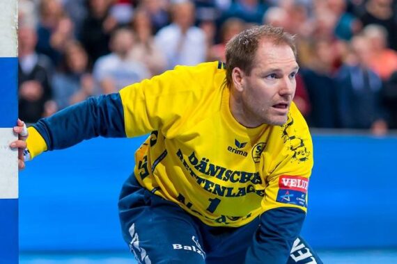 Ein männlicher Handballtorwart in einem gelben Trikot und einer blauen Hose steht aufmerksam da, hält sich mit einer Hand am Torpfosten fest und ist bereit, den Ball abzuwehren. Eine verschwommene Menschenmenge schaut im Hintergrund zu.