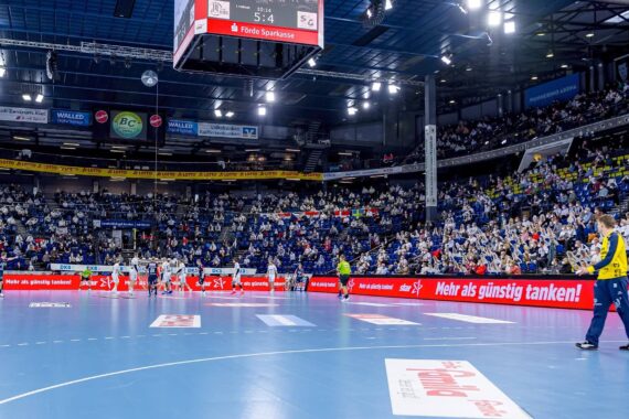 Weite Sicht auf eine große, mit Zuschauern gefüllte Handballhalle mit Spielern auf dem blauen Spielfeld und einer digitalen Anzeigetafel über dem Spielfeld, die ein Ergebnis von 5:4 anzeigt. Helle Beleuchtung und Werbebanner sind rund um die Halle zu sehen.