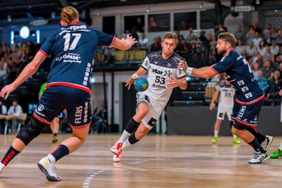 Ein Handballspieler in einem weißen Trikot dribbelt den Ball, während zwei Gegner in dunkelblauen Trikots versuchen, ihn auf einem Hallenplatz zu blockieren, während im Hintergrund eine Menschenmenge zusieht.