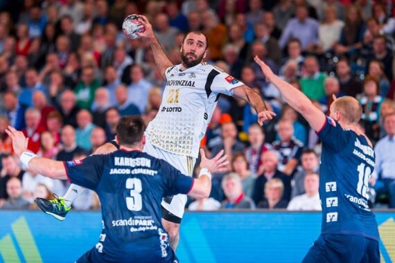 Ein Handballspieler in einem weißen Trikot springt, um den Ball zu werfen, während zwei Verteidiger in blauen Trikots versuchen, ihn zu blockieren. Das Spiel findet in einer überfüllten Halle mit Zuschauern im Hintergrund statt.