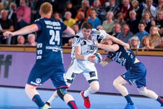 Ein Handballspieler in einem weißen Trikot versucht, während eines intensiven Hallenspiels an zwei Verteidigern in dunkelblauen Trikots vorbeizukommen, während im Hintergrund eine Zuschauermenge zusieht.