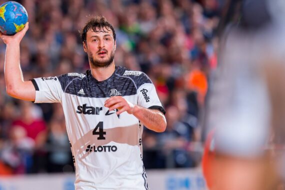 Ein männlicher Handballspieler in einem weiß-schwarzen Trikot mit der Nummer 4 hält einen blauen Ball und bereitet sich auf den Wurf vor, während eines Spiels mit einer verschwommenen Menschenmenge im Hintergrund.