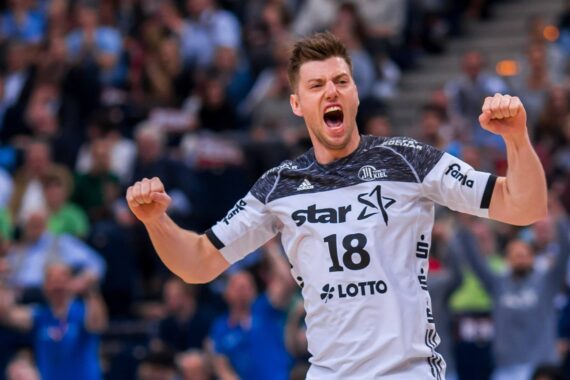 Ein männlicher Handballspieler in einem weiß-schwarzen Trikot mit der Nummer 18 feiert leidenschaftlich auf dem Spielfeld, mit erhobenen Fäusten und offenem Mund, mit einer verschwommenen Menschenmenge im Hintergrund.
