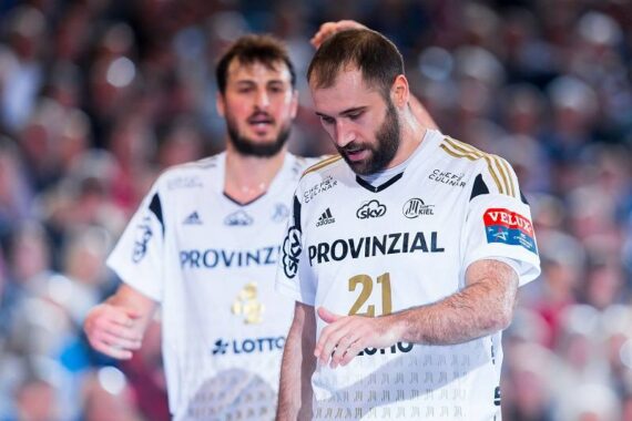 Zwei männliche Handballspieler in weißen Trikots mit goldenen Akzenten stehen auf einem Spielfeld. Der vordere Spieler schaut konzentriert nach unten, während der Spieler hinter ihm nach vorne schaut. Im Hintergrund ist eine verschwommene Menschenmenge zu sehen.