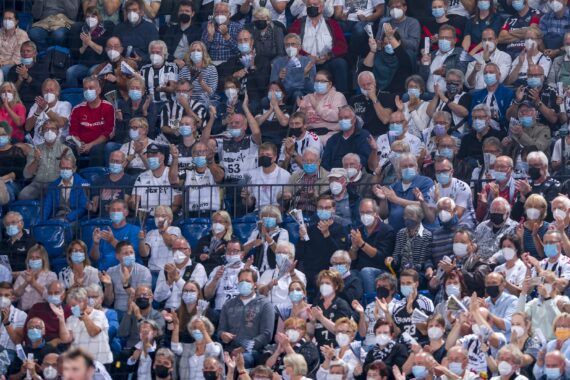 Eine große Menschenmenge mit Gesichtsmasken sitzt in einem Stadion dicht beieinander und verfolgt eine Veranstaltung. Viele klatschen, einige stehen, und die meisten sind leger gekleidet.