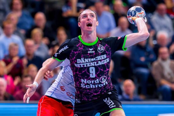 Ein männlicher Handballspieler in einem schwarz-lila Trikot mit der Nummer 9 bereitet sich auf den Wurf vor, während ein rot-weiß gekleideter Verteidiger versucht, ihn abzuwehren. Die Aktion findet vor einer verschwommenen Menschenmenge statt.