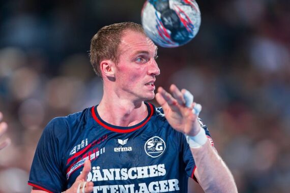 Ein männlicher Handballspieler in einem marineblauen Trikot konzentriert sich während eines Spiels auf einen mehrfarbigen Handball, der in der Nähe seines Gesichts schwebt. Der Hintergrund ist unscharf, um die Action und die Intensität des Moments hervorzuheben.