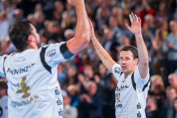 Zwei männliche Handballspieler in weißen Trikots feiern mit erhobenen Armen vor jubelnden Zuschauern im Hintergrund. Der Spieler auf der rechten Seite blickt nach vorne, während der andere von der Kamera wegschaut.