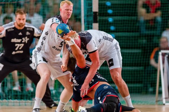 Zwei Handballspieler in weißen Trikots verteidigen sich gegen einen Spieler in einem dunkelblauen Trikot, der mit dem Ball in der Hand zu Boden fällt; im Hintergrund steht ein Torwart in der Nähe des Tors.