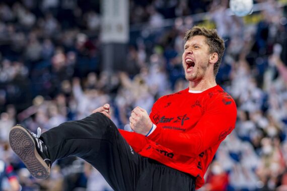 Ein männlicher Handballspieler in einem roten Trikot jubelt leidenschaftlich, ballt die Fäuste und schreit vor Begeisterung auf dem Spielfeld, während im Hintergrund eine verschwommene Menge jubelt.