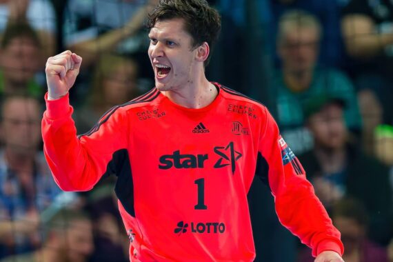 Ein männlicher Handballspieler in einem leuchtend roten Trikot mit der Nummer 1 jubelt während eines Spiels mit geballter Faust und entschlossenem Gesichtsausdruck, während im Hintergrund eine unscharfe Menschenmenge zu sehen ist.