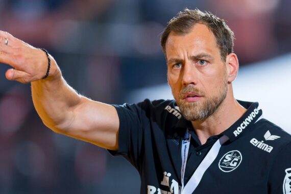 Ein Mann, der ein schwarzes Sporthemd mit Logos trägt, gestikuliert mit dem ausgestreckten rechten Arm und schaut konzentriert. Er hat kurze Haare, einen gestutzten Bart und trägt ein Schlüsselband um den Hals. Der Hintergrund ist unscharf.