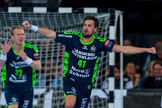 Ein Handballspieler in einem dunkelblauen und neongrünen Trikot jubelt mit ausgestreckten Armen auf dem Spielfeld, während ein anderer Mitspieler hinter ihm klatscht, beide vor dem Tornetz.