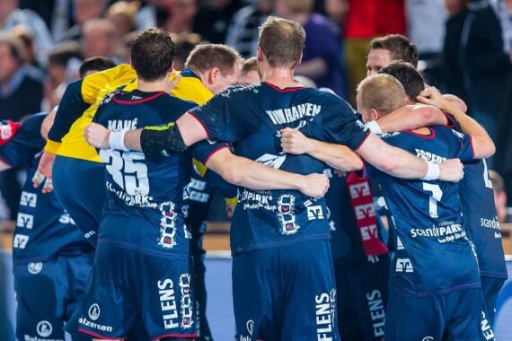 Eine Gruppe von männlichen Handballspielern in blauen Trikots steht während eines Spiels auf dem Spielfeld zusammen, die Arme umeinander gelegt und von der Kamera abgewandt.