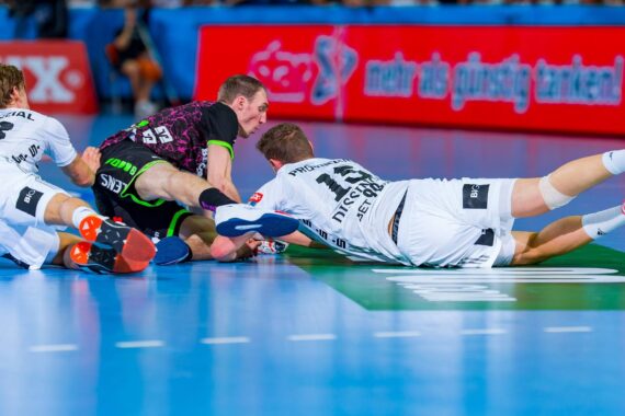 Drei männliche Handballspieler kämpfen auf einem blauen Hallenplatz um den Ball. Zwei Spieler in weißen Trikots rutschen auf einen Spieler in einem schwarz-grünen Trikot zu, alle strecken sich intensiv nach vorne.