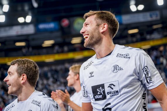 Zwei männliche Sportler in weißen Trikots lächeln und feiern in einer Halle, wahrscheinlich nach einem Sportspiel. Der Hintergrund zeigt eine jubelnde Menge und helle Stadionlichter.