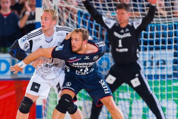 Zwei männliche Handballspieler, einer in einem weißen und einer in einem dunkelblauen Trikot, spielen in der Nähe des Tores um die Wette, während ein Torwart in Schwarz mit erhobenen Armen vor dem Tor steht.