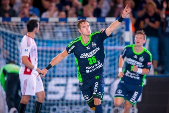 Ein männlicher Handballspieler in einem dunkelblauen und grünen Trikot jubelt mit einem erhobenen Arm, der nach oben zeigt, während im Hintergrund andere Spieler und ein Netztor zu sehen sind.