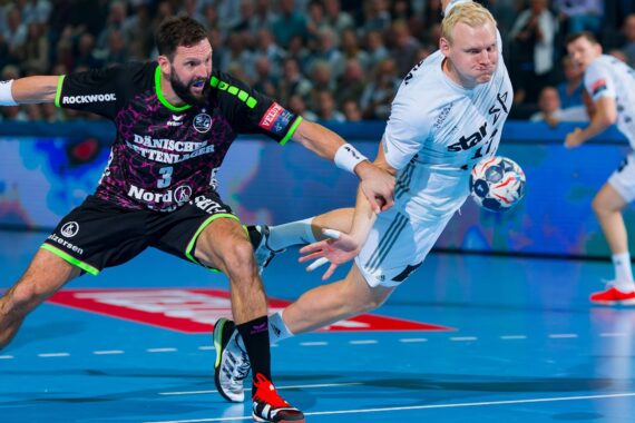 Zwei männliche Handballspieler kämpfen auf einem Hallenplatz um den Ball; der eine in einem schwarz-grünen Trikot greift nach dem anderen in einem weißen Trikot, der mit dem Ball in der Hand nach vorne springt.