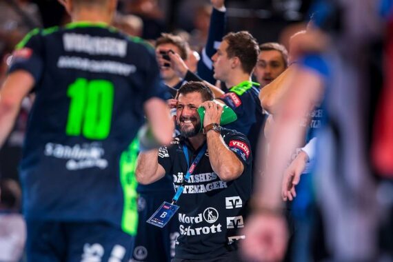 Ein Handballtrainer in einem marineblauen Hemd hält sich den Kopf und schreit vor Aufregung oder Frustration, umgeben von Spielern und Betreuern, die während eines Spiels in Aktion sind. Die Szene ist energiegeladen, mit unscharfen Bewegungen und bunten Uniformen.