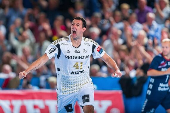 Ein männlicher Handballspieler, der ein weißes Trikot mit der Nummer 41 trägt, schreit auf dem Spielfeld vor Freude. Der Hintergrund zeigt eine unscharfe Menge und andere Spieler in blauen und roten Trikots.