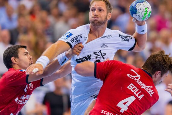 Ein männlicher Handballspieler in einem weißen Trikot springt mit dem Ball, während zwei Gegner in roten Trikots während eines intensiven Spiels eng verteidigen. Im Hintergrund ist eine unscharfe Menschenmenge zu sehen.