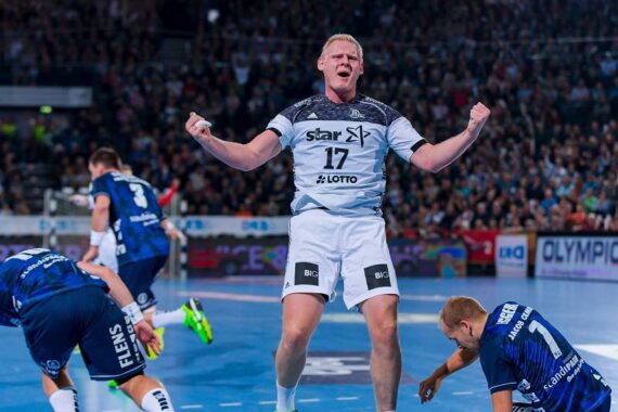 Ein Handballspieler in einem weiß-grauen Trikot mit der Nummer 17 feiert energisch mit geballten Fäusten auf einem Hallenplatz, während gegnerische Spieler in blauen Trikots um ihn herum reagieren. Im Hintergrund sind Zuschauer zu sehen.