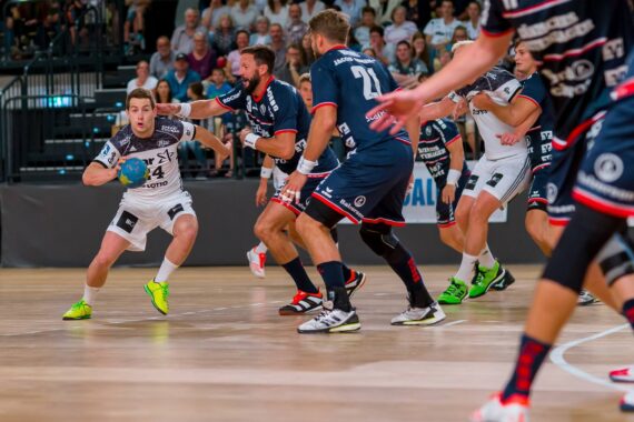 Ein Handballspieler in einem weißen Trikot läuft mit dem Ball, während er von mehreren Spielern in blauen Trikots auf einem Hallenplatz eng verteidigt wird, während im Hintergrund Zuschauer zusehen.