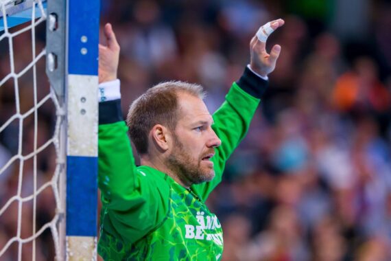 Ein Handballtorwart in einem grünen Trikot mit erhobenen Armen steht während eines Spiels wachsam in der Nähe des Torpfostens, mit einer verschwommenen Menschenmenge im Hintergrund.