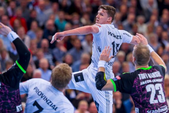 Ein männlicher Handballspieler in einem weißen Trikot springt in die Luft, um den Ball zu werfen, während zwei gegnerische Spieler in schwarzen Trikots mit neongrünen Akzenten versuchen, ihn während eines Spiels zu blockieren. Die Zuschauer sind im Hintergrund unscharf zu erkennen.