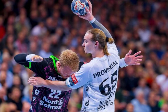 Zwei männliche Handballspieler liefern sich einen intensiven Wettkampf; der eine in einem weißen Trikot hält den Ball hoch, während der andere in einem dunklen Trikot sich in die Defensive drängt. Eine unscharfe Menge schaut im Hintergrund zu.