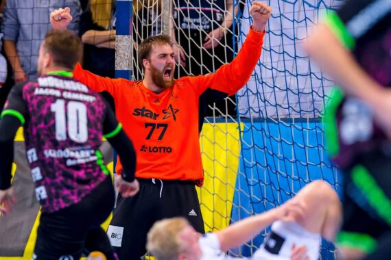 Ein Handballtorwart in einem orangefarbenen Trikot jubelt mit erhobenen Fäusten und aufgerissenem Mund, während Spieler beider Mannschaften auf dem Spielfeld vor ihm reagieren. Im Hintergrund sind ein Netz und verschwommene Zuschauer zu sehen.