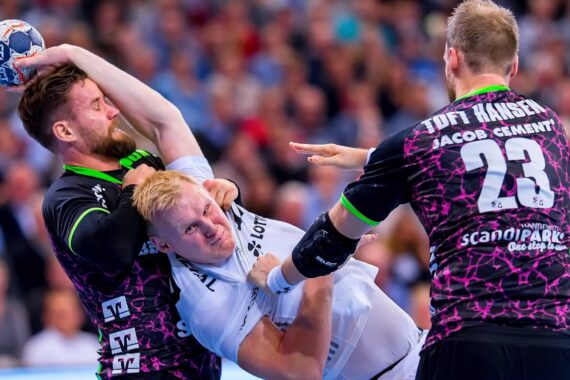Ein Handballspieler in einem weißen Trikot wird während eines Spiels von einem Verteidiger in einem schwarzen Trikot fest am Hals und am Oberkörper gepackt, während sich ein anderer Spieler in Schwarz nähert. Die Szene ist intensiv und körperlich.
