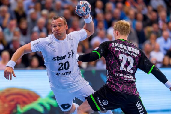 Ein Handballspieler in einem weißen Trikot springt, um den Ball zu werfen, während ein Abwehrspieler in einem schwarz-lila Trikot versucht, ihn zu blockieren. Das Geschehen ist intensiv, und im Hintergrund schaut eine Menschenmenge zu.