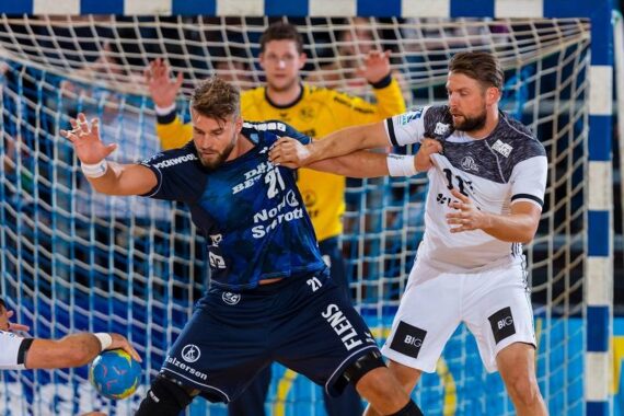 Zwei männliche Handballspieler, einer in einem blauen und der andere in einem weißen Trikot, liefern sich in der Nähe des Tores einen intensiven Wettkampf, während im Hintergrund ein Torwart in Gelb bereitsteht. Der Spieler in Blau hält den Ball, der von dem Spieler in Weiß eng verteidigt wird.
