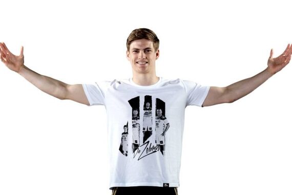 Ein junger Mann, der ein weißes grafisches T-Shirt trägt, steht mit ausgestreckten Armen und lächelt vor einem schlichten weißen Hintergrund.