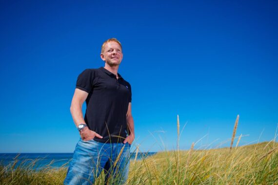 Ein Mann in schwarzem Polohemd und Jeans steht im hohen Gras in der Nähe des Meeres unter einem klaren blauen Himmel, die Hände in den Taschen und mit entspannter Miene.