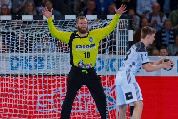 Ein männlicher Handballtorwart in einem gelben Trikot steht mit erhobenen Armen vor dem Tor und bereitet sich auf die Abwehr vor. Ein Spieler in einem weiß-schwarzen Trikot hält den Ball in der Nähe. Die Zuschauer schauen von der Tribüne im Hintergrund zu.