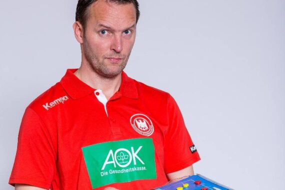 Ein Mann in einem roten Sporthemd mit Logos hält eine blaue Taktiktafel mit farbigen Magneten und blickt mit einem leichten Lächeln in die Kamera.