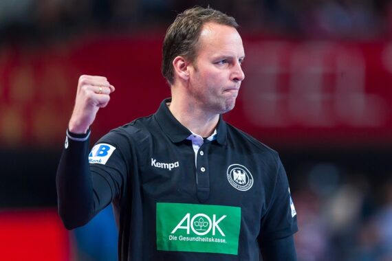 Ein Handballtrainer in einem schwarzen Hemd mit dem Logo der deutschen Mannschaft hebt während eines Spiels entschlossen die Faust, während im Hintergrund eine unscharfe Menschenmenge zu sehen ist.