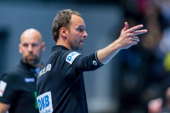 Ein Mann in einer schwarzen Jacke gestikuliert mit seiner rechten Hand, als wolle er Anweisungen geben. Ein anderer Mann in einem ähnlichen Outfit steht etwas hinter ihm. Beide befinden sich in einer Sporthalle.